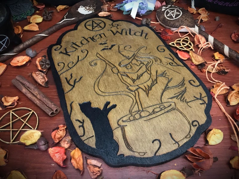 Op de afbeelding: Een houten bord met een zwarte rand, waarop een heks een ketel roert met een kat die toekijkt. Het bord heeft de tekst "Kitchen Witch" met een pentagram symbool boven de tekst.