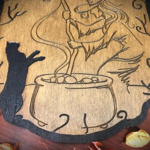 Op de afbeelding: Een houten wanddecoratie met een zwart katten silhouet, een heks die een ketel roert en ingewikkelde ontwerpen. De achtergrond is van lichtbruin hout.