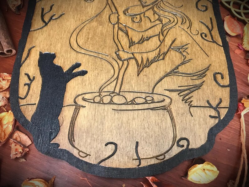 Op de afbeelding: Een houten wanddecoratie met een zwart katten silhouet, een heks die een ketel roert en ingewikkelde ontwerpen. De achtergrond is van lichtbruin hout.