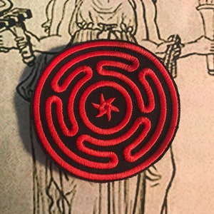 Può includere: Una toppa ricamata rossa e nera con un design circolare che presenta una stella a cinque punte al centro e cerchi concentrici attorno.