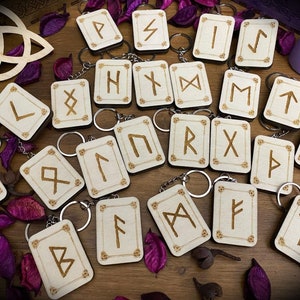 Könnte beinhalten: Ein Set mit 18 Holzschlüsselanhängern mit den Runen des älteren Futhark-Alphabets. Jede Rune ist in ein kleines, rechteckiges Stück Holz mit hellbrauner Oberfläche graviert. Die Schlüsselanhänger sind in einem zufälligen Muster auf einer Holzoberfläche angeordnet.