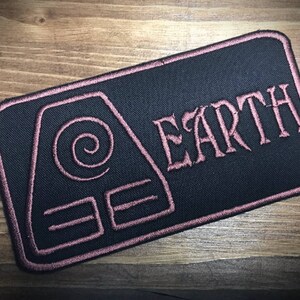 Może przedstawiać: Czarna haftowana naszywka z różowym obramowaniem. Naszywka przedstawia stylizowany symbol ziemi i napis "EARTH" różowymi literami.