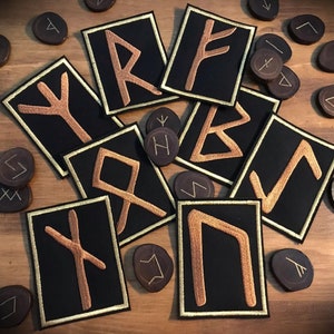 Peut inclure: Neuf patchs en tissu noir avec une bordure dorée, chacun représentant un symbole de rune brodé différent.