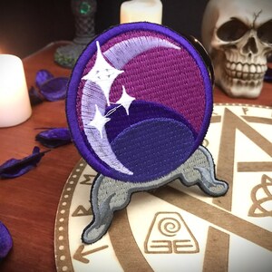 Pode incluir: Patch bordado roxo e azul de uma lua crescente com estrelas brancas, em um suporte cinza, em uma superfície de madeira com um design circular.