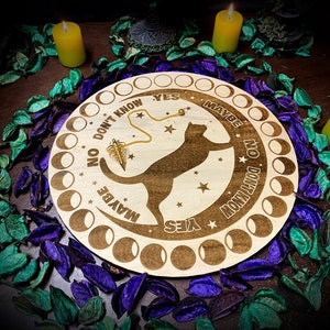 Könnte beinhalten: Ein Ouija-Brett aus Holz mit einem schwarzen Katzenmotiv und den Worten "Ja", "Vielleicht", "Nein" und "Weiß nicht" am Rand. Ein goldenes Pendel hängt in der Mitte des Bretts.
