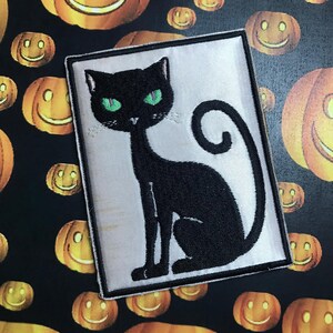 Könnte beinhalten: Eine schwarze Katze mit grünen Augen sitzt auf weißem Hintergrund. Die Katze ist auf einem Patch gestickt.