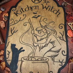 Op de afbeelding: Een houten bord met een zwarte rand en een pentagram bovenaan. Het bord zegt "Kitchen Witch" en toont een heks die een ketel roert met een zwarte kat die toekijkt.