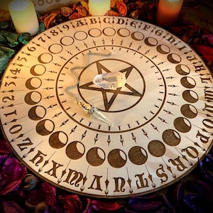 Könnte beinhalten: Ein Ouija-Brett aus Holz mit einem Pentagramm-Design und einem Kristallpendel. Das Brett zeigt die Zahlen 1-31, die Buchstaben A-Z und Mondphasen. Das Brett ist von Kerzen und violetten und grünen Blütenblättern umgeben.