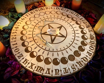 Może przedstawiać: Drewniana plansza Ouija z wzorem pentagramu i kryształowym wahadłem. Plansza zawiera liczby od 1 do 31, litery od A do Z i fazy księżyca. Plansza jest otoczona świecami i fioletowymi i zielonymi płatkami kwiatów.