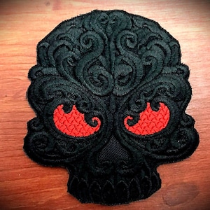 Peut inclure: Patch brodé noir en forme de crâne avec des détails complexes et des yeux rouges en forme de flammes.