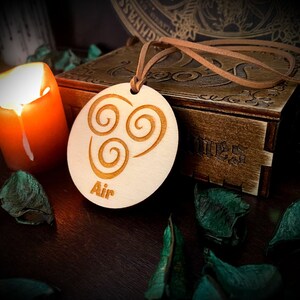 Könnte beinhalten: Ein helles Holzanhänger mit dem Wort "Air" und einem Wirbelsymbol, das an einer braunen Kordel hängt. Das Symbol und der Text sind in das Holz geätzt. Eine brennende orangefarbene Kerze und eine Holzkiste befinden sich im Hintergrund.