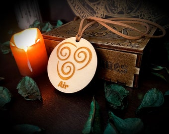 May include: A wooden pendant with the word "Air" and a swirling symbol, hanging from a brown cord. The pendant is light-colored, with the symbol and text etched into the wood. A lit orange candle and a wooden box are in the background.