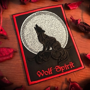 Puede incluir: Parche bordado negro con borde rojo. El parche presenta un lobo aullando a una luna blanca con un diseño de remolino blanco y negro. El texto "Wolf Spirit" está bordado en rojo debajo del lobo.