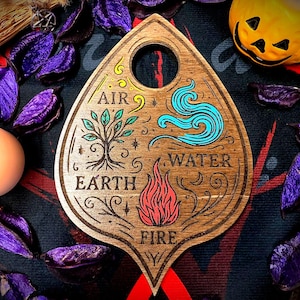 Könnte beinhalten: Eine Holzplanchette mit einem Loch oben, mit eingravierten Symbolen, die die Elemente darstellen: Luft, Erde, Wasser und Feuer. Die Planchette ist von lila Potpourri, einem Kürbis und einer weißen Kerze umgeben.