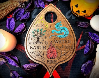 May include: A wooden planchette with a hole at the top, featuring engraved symbols representing the elements: Air, Earth, Water, and Fire. The planchette is surrounded by purple potpourri, a pumpkin, and a white candle.