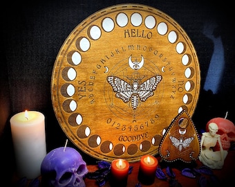 Może przedstawiać: Drewniana plansza Ouija z motywem ćmy, fazami księżyca i literami alfabetu. Plansza jest otoczona świecami i małą drewnianą planszetką.
