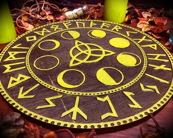 Puede incluir: Un círculo de madera con un diseño pintado en amarillo de un nudo celta y seis fases de la luna. El círculo está rodeado por un anillo de runas amarillas.