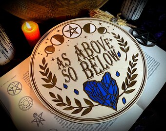 Puede incluir: Una placa redonda de madera con el texto "As Above So Below" en letras doradas. La placa presenta un pentagrama, una luna creciente y un corazón de cristal azul.