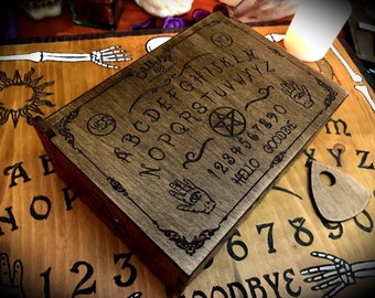 Puede incluir: Una tabla de Ouija de madera con una mancha marrón oscura. La tabla tiene el alfabeto, los números del 1 al 9 y el 0, las palabras "hola", "adiós" y "sí" impresas en ella. La tabla está cerrada y tiene un pentagrama y dos manos con un ojo en el centro.