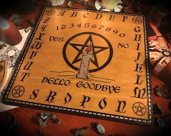 Może przedstawiać: Drewniana plansza Ouija z pentagramem, zapalonym świecą i literami od A do Z, liczbami od 1 do 0 oraz słowami "tak", "nie", "cześć" i "do widzenia" nadrukowanymi na niej.
