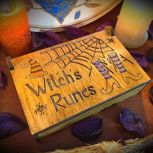 Puede incluir: Caja de madera con un diseño de sombrero de bruja, telaraña y botas de bruja. La caja está etiquetada "Witch's Runes".