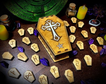 Puede incluir: Caja de madera en forma de ataúd con un diseño de cruz blanca y estrella. La caja está rodeada de runas de madera sobre una superficie de madera oscura.