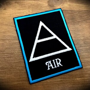 Peut inclure: Un patch brodé noir avec un triangle blanc et le mot "AIR" en lettres blanches. Le patch a une bordure bleu clair.