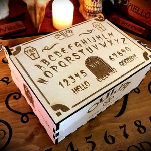 Op de afbeelding: Een houten doos met een Ouija-bordontwerp. De doos is lichtbruin en heeft een wit interieur. Het Ouija-bordontwerp bevat de letters A tot en met Z, de cijfers 1 tot en met 9 en 0, de woorden "YES", "NO", "GOODBYE", "HELLO" en "RIP".