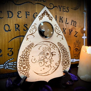 Può includere: Una planchette di tavola Ouija in legno con un design a mezzaluna e floreale. La planchette ha una lente di vetro trasparente al centro. La planchette è appoggiata su una tavola Ouija in legno con le lettere dell'alfabeto, i numeri da 1 a 9 e le parole "sì" e "no".