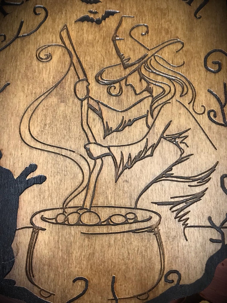Op de afbeelding: Een houten bord met een zwart-witte illustratie van een heks die een ketel roert. De heks draagt een punthoed en heeft lang haar. De ketel is gevuld met bubbels. Het bord heeft een rustieke, vintage uitstraling.