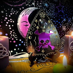 Może przedstawiać: Czarna drewniana plansza Ouija z purpurowym wzorem czaszki i skrzyżowanych kości, kryształową kulą i złotymi akcentami. Plansza stoi na stojaku i jest otoczona fioletowymi świecami z celtyckimi wzorami węzłów.
