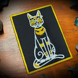 May include: A black and white embroidered patch of a cat with gold accents. The cat is sitting with its tail curled around its legs. The patch has a yellow border.