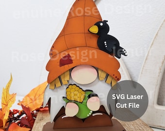 Pode incluir: Uma figura de gnomo de madeira com um pássaro preto empoleirado em seu chapéu. O gnomo está segurando uma espiga de milho verde. O gnomo é pintado em cores outonais, incluindo laranja, marrom e amarelo. O gnomo está em pé sobre uma base marrom. Arquivo de corte a laser SVG.