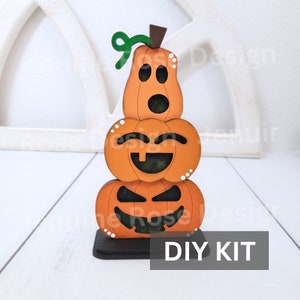May include: A DIY kit for a stacked pumpkin decoration. The kit includes three wooden pumpkins with different facial expressions, a green stem, and a brown base. The pumpkins are orange with black facial features and white dots.