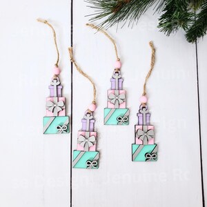May include: Four wooden Christmas ornaments shaped like stacked gift boxes. Each ornament is decorated with a pink, purple, and teal color scheme and a silver bow. The ornaments are strung on twine with a wooden bead at the top.