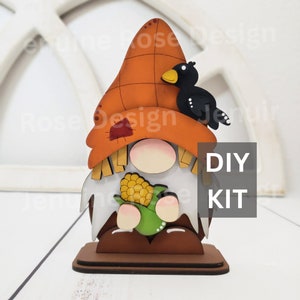 May include: DIY kit for a wooden gnome figurine with a black bird perched on its hat, holding a green corn cob. The gnome is wearing a brown hat with a red patch and a white beard. The base is brown wood.