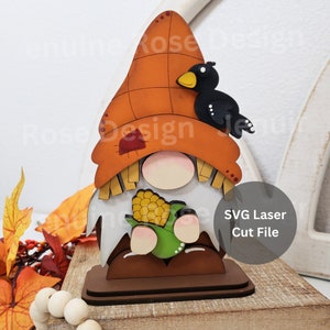 May include: A wooden gnome figurine with a black bird perched on its hat. The gnome is holding a green corn cob. The gnome is painted in autumnal colours, including orange, brown, and yellow. The gnome is standing on a brown base. SVG Laser Cut File.