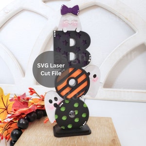 May include: A wooden Halloween decoration featuring the letters "BOO" stacked on top of each other. Each letter is decorated with a different pattern, and a small ghost with a purple bow sits on top of the "B".