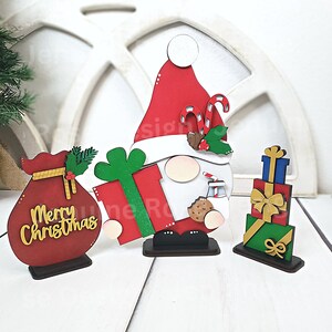 May include: A wooden Christmas gnome figurine holding a red and green gift box. The gnome is wearing a red hat with white trim and a white beard. The gnome is standing on a wooden base. The gnome is surrounded by a red and gold bag with the words "Merry Christmas" and a stack of wrapped gifts.