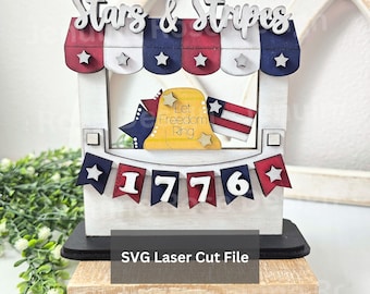 May include: A tiered tray decor piece featuring a wooden stand with a red, white, and blue striped awning, the text Stars & Stripes, a yellow bell with the text Let Freedom Ring, and a banner displaying the year 1776 with star accents.