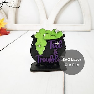 May include: A black wooden cauldron with green and white bubbling potion and the text "Toil & Trouble" in purple. SVG Laser Cut File.