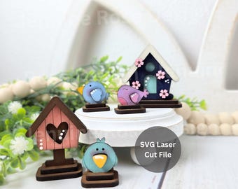 May include: A set of laser-cut wooden decorations featuring two birdhouses and two small birds. One birdhouse is pink with a heart cutout, and the other is dark blue with white trim and pink flowers. The birds are blue and purple with yellow beaks.
