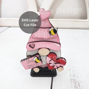 Op de afbeelding: Een roze en zwarte houten gnome die een bordje vasthoudt met de tekst "Bee Mine" en een hartvormige doos met een strik. De gnome heeft een roze hoed met een hartje erop en een bij aan de zijkant.
