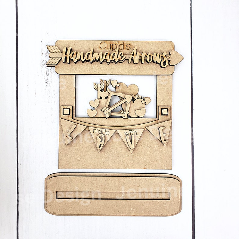 Op de afbeelding: Een houten ambacht met de tekst "Cupid's Handmade Arrows". Het ontwerp omvat een uitsparing van Cupido met pijlen en harten, en een banner met de tekst "made with love". Het ambacht is onafgewerkt en klaar om te worden geverfd of versierd.