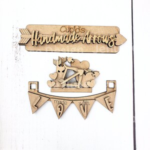 Op de afbeelding: Houten Valentijnsdagdecoraties. De set bevat een bord met de tekst "Cupid's Handmade Arrows", een Cupido met pijlen en harten, en een banner met "made with love".