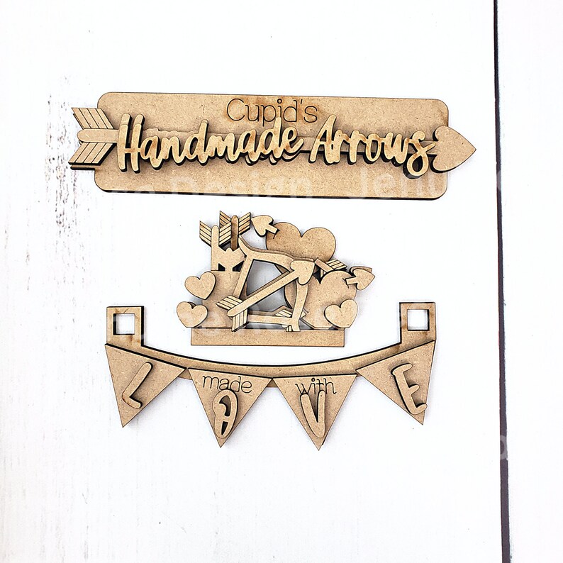 Op de afbeelding: Houten Valentijnsdagdecoraties. De set bevat een bord met de tekst "Cupid's Handmade Arrows", een Cupido met pijlen en harten, en een banner met "made with love".