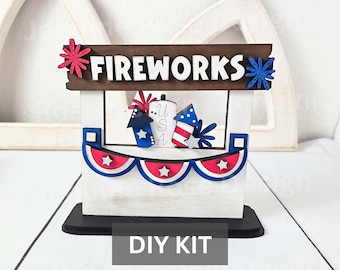 May include: A wooden tabletop display featuring a brown sign with the white text FIREWORKS, accented by red and blue starbursts. The stand includes a white base with a red, white, and blue bunting, and a central cutout displaying USA text.