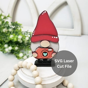 May include: A wooden gnome figurine with a red hat, white beard, and a red camera with a blue heart. The gnome is standing on a black base. The text "SVG Laser Cut File" is visible in the background.