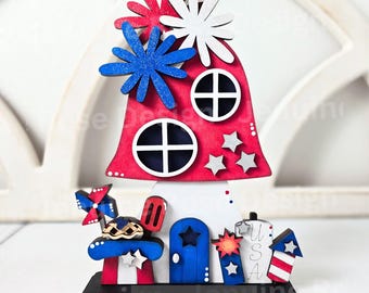 May include: A wooden DIY kit featuring a red mushroom-shaped house with white and blue star accents, topped with red, white, and blue glittery flower shapes. The base includes a blue door, a small pie, a pinwheel, and a sign with the text USA.
