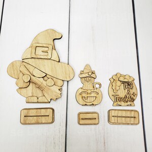May include: Wooden cutout set of three Halloween characters: a witch holding a broom, a cat in a witch hat, and a cauldron with the text "Toil & Trouble".